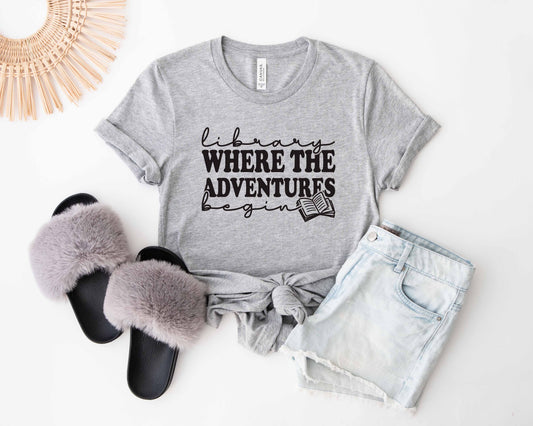 An athletic heather color shirt with the saying "Library where the adventures begin"