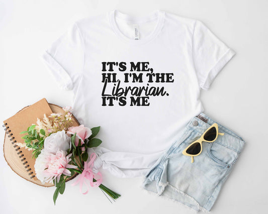 A white color shirt with the saying "It's me hi im the librarian its me"