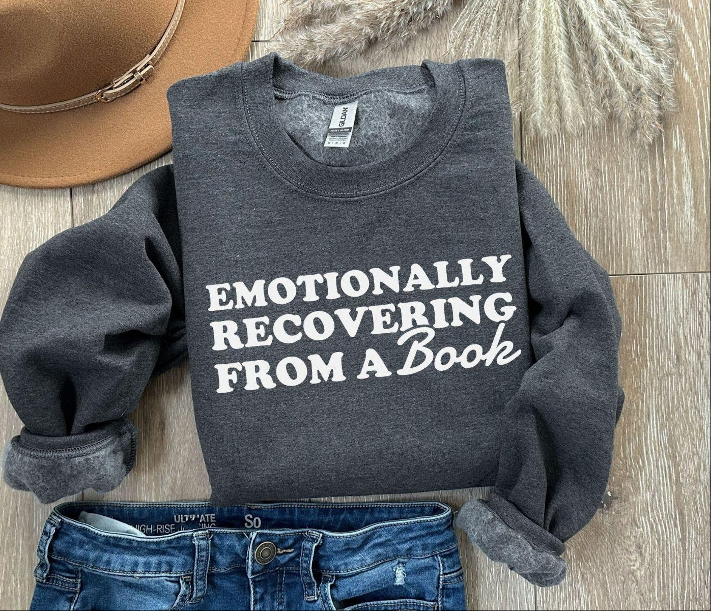 Dark gray sweatshirt with 'Emotionally Recovering from a Book' text, paired with jeans and a hat on a wooden surface.