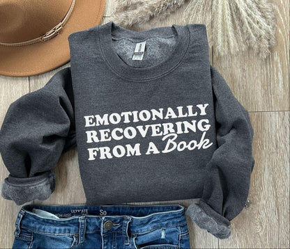 Dark gray sweatshirt with 'Emotionally Recovering from a Book' text, paired with jeans and a hat on a wooden surface.
