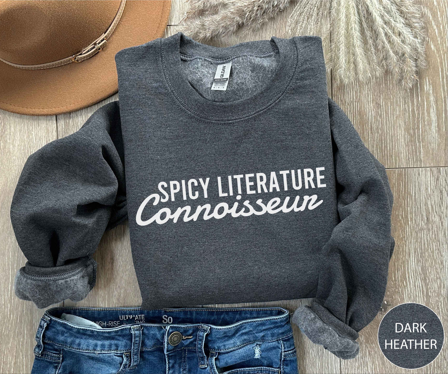 Dark Heather sweatshirt with 'Spicy Literature Connoisseur' text, placed on a wooden surface with jeans and a hat.