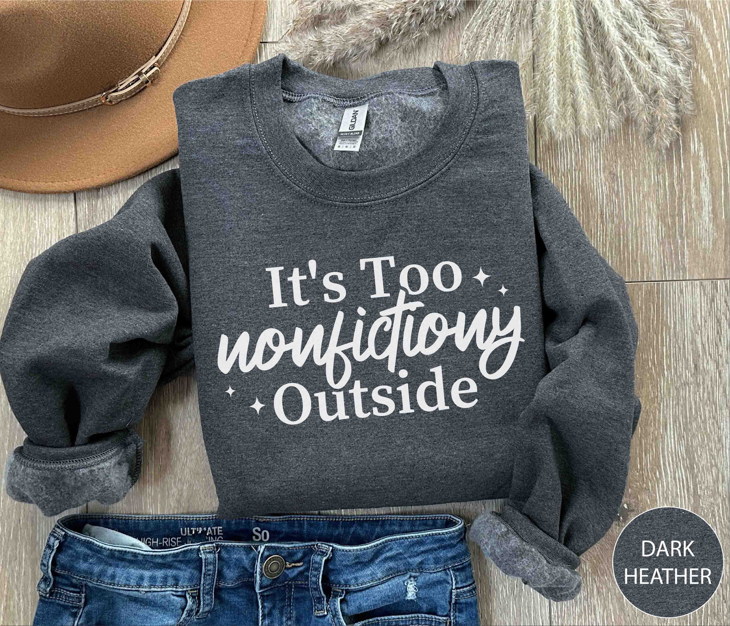 Dark Heather sweatshirt with 'It's Too Nonfictiony Outside' text on a wooden surface with jeans and a hat.