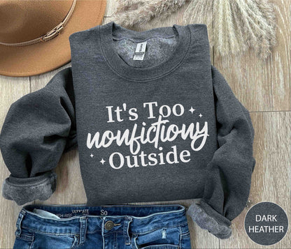 Dark Heather sweatshirt with 'It's Too Nonfictiony Outside' text on a wooden surface with jeans and a hat.