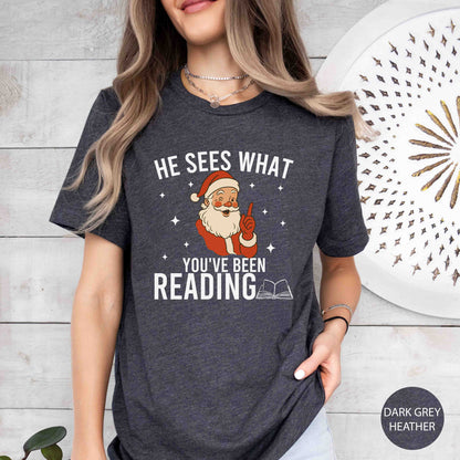 Person wearing a dark grey t-shirt with a Santa Claus graphic and text, standing against a light wooden wall.