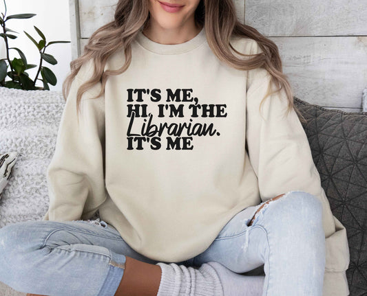 A sand color sweatshirt with the saying "Its me hi i'm the librarian it's me"