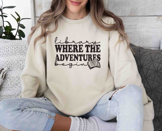 A sand color sweatshirt with the saying "Library where the adventures begin"