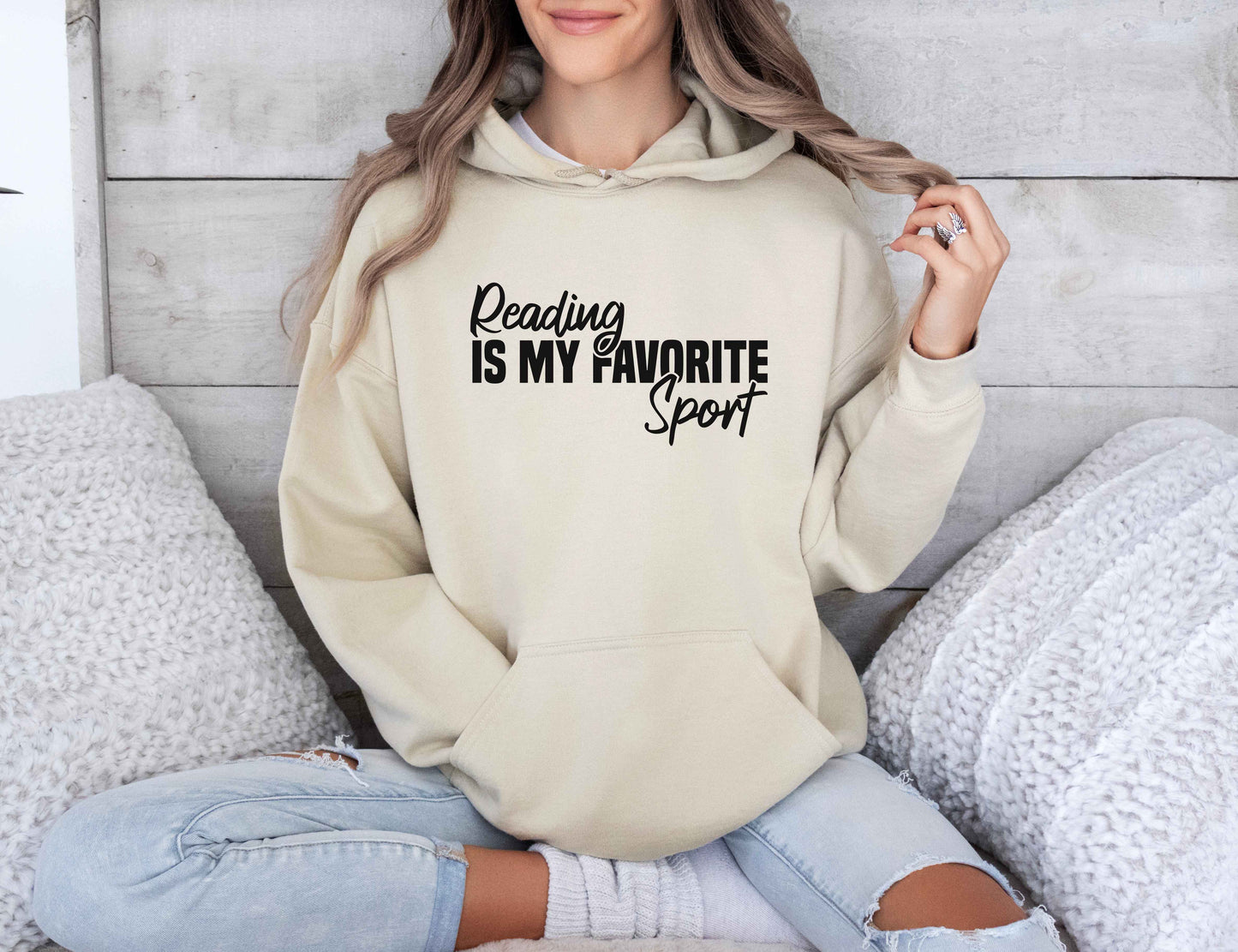 Person wearing a sand color hoodie with 'Reading is my favorite sport' text, sitting on a couch.