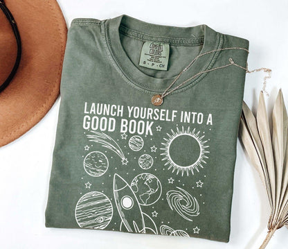 A comfort colors® shirt with the saying "launch yourself into a good book reading is out of this world"