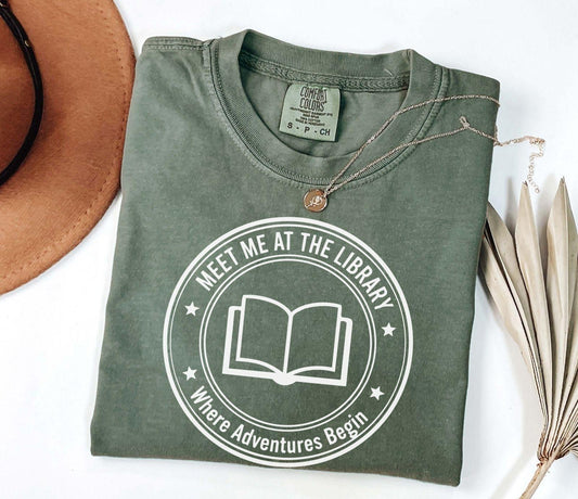 A comfort colors® shirt with the saying "meet me at the library where adventure begin"