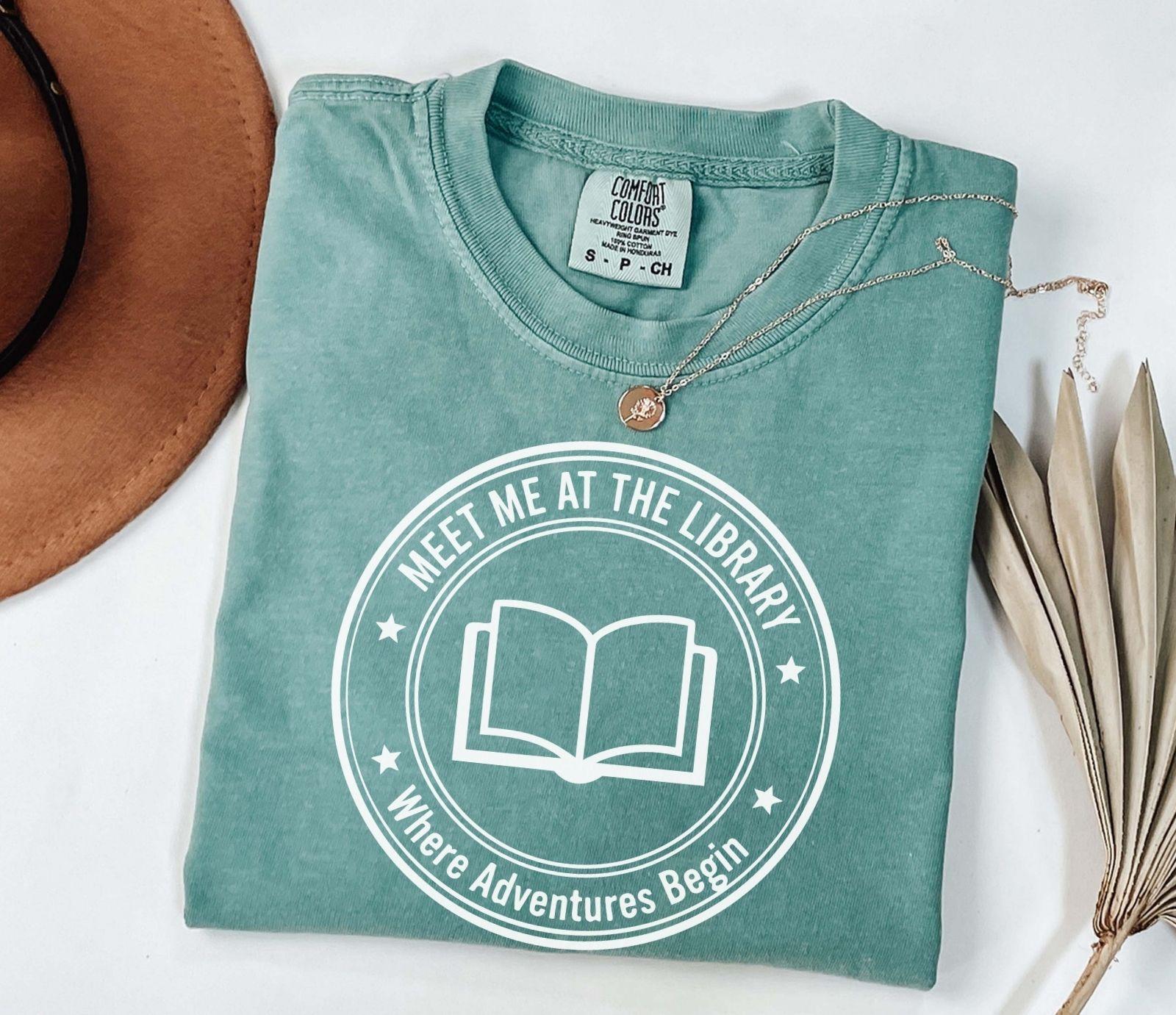 A comfort colors® shirt with the saying "meet me at the library where adventure begin"
