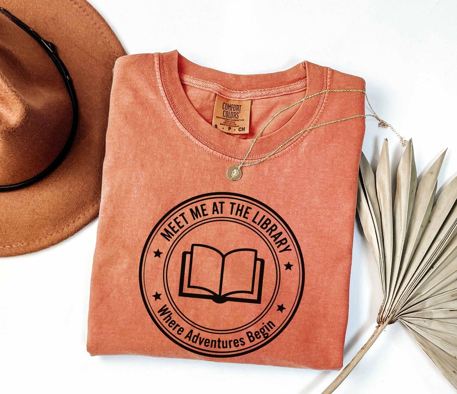 A comfort colors® shirt with the saying "meet me at the library where adventure begin"