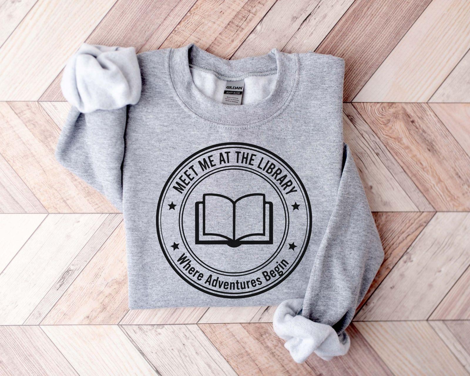 A Gildan 18000 sweatshirt with the saying "meet me at the library where adventures begin"
