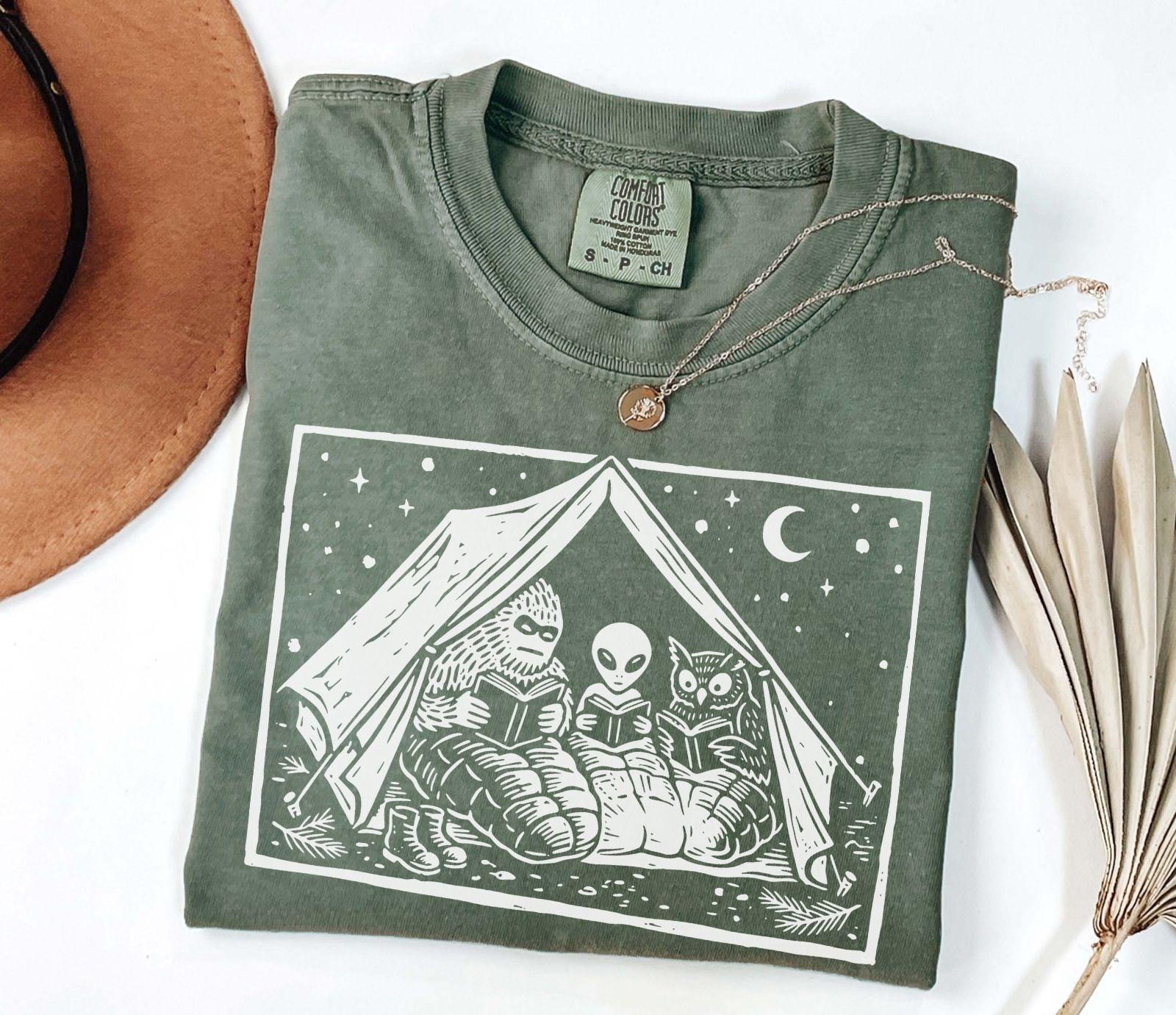 A comfort colors® shirt with the saying "Mothman Bigfoot and Horned reading a book"
