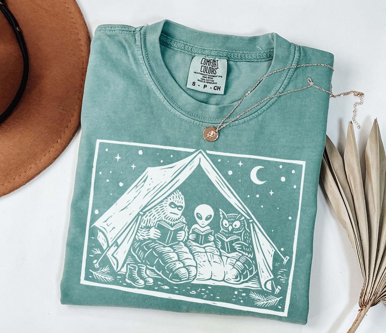 A comfort colors® shirt with the saying "Mothman Bigfoot and Horned reading a book"