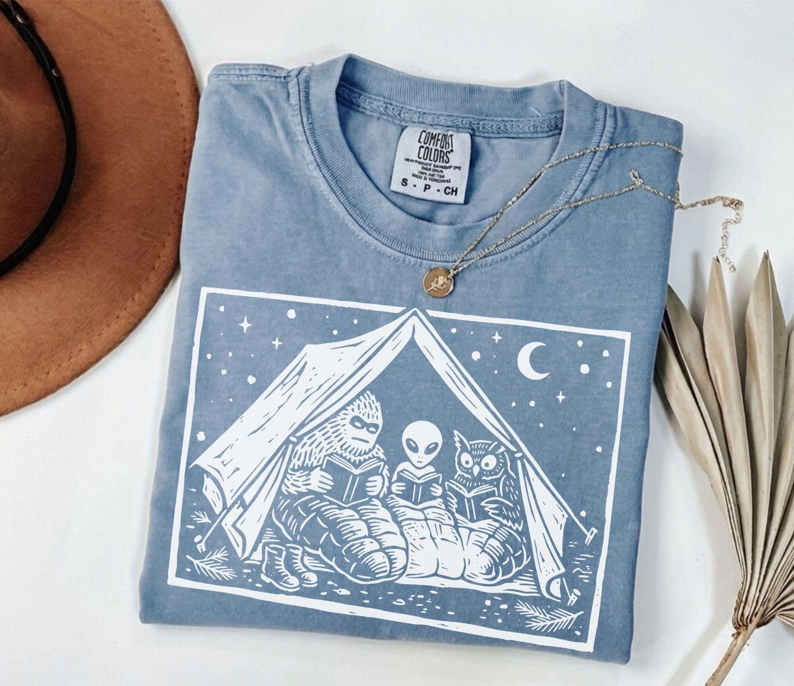 A comfort colors® shirt with the saying "Mothman Bigfoot and Horned reading a book"