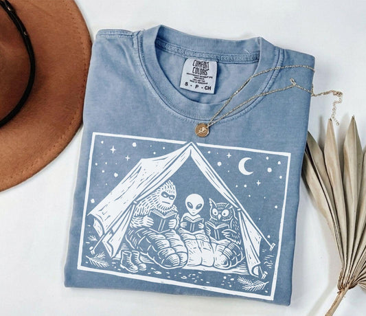A comfort colors® shirt with the saying "Mothman Bigfoot and Horned reading a book"