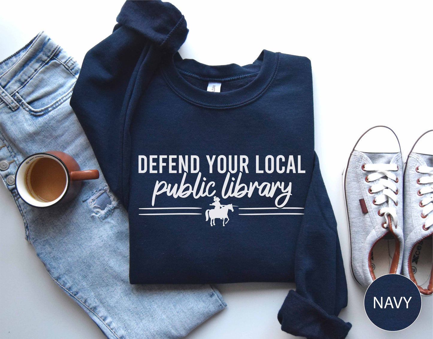 Navy sweatshirt with 'Defend Your Local Public Library' text, placed on a white surface with jeans, a mug, and shoes.