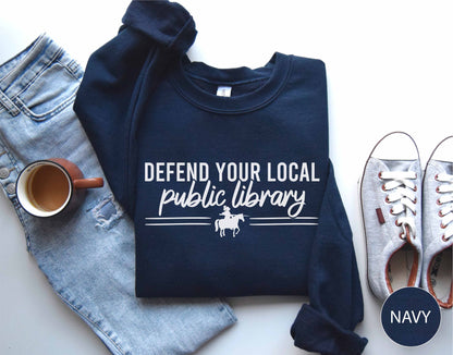 Navy sweatshirt with 'Defend Your Local Public Library' text, placed on a white surface with jeans, a mug, and shoes.