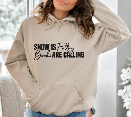A sand color hoodie with the saying "Snow is falling books are calling"